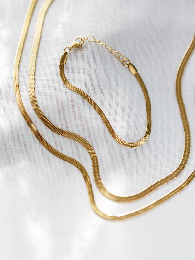 Set aus zwei flachen Schlangenketten in Gold mit passendem Armband in Gold auf hellem Stoff